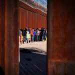 Work Permit Crackdown Shocks Asylum System A line of people waiting near a border wall with barbed wire