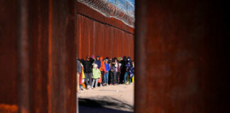 A line of people waiting near a border wall with barbed wire