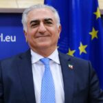 Smiling man in a suit with a blue tie, standing in front of an EU flag