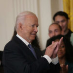 Biden Officials QUIETLY Alter Stroke Risk Warnings A smiling man in a suit clapping during a formal event with a crowd in the background