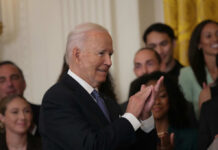 A smiling man in a suit clapping during a formal event with a crowd in the background