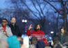 A speaker in a red coat addresses a crowd at a political rally during dusk