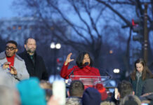 A speaker in a red coat addresses a crowd at a political rally during dusk