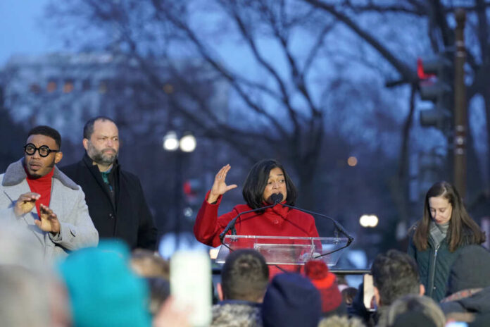 A speaker in a red coat addresses a crowd at a political rally during dusk