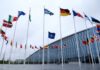 Flags of various countries in front of a modern building under a cloudy sky