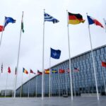Trump’s SHOCKING Threat to NATO: Act or Collapse Flags of various countries in front of a modern building under a cloudy sky