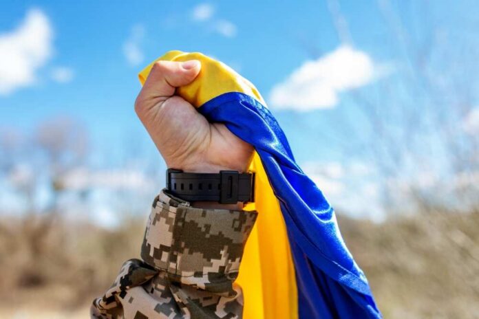 shutterstock_2303336965.jpg A soldier holding a blue and yellow flag against a bright sky