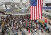 Crowd of travelers waiting in line at an airport security checkpoint under an American flag