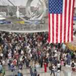 Crowd of travelers waiting in line at an airport security checkpoint under an American flag
