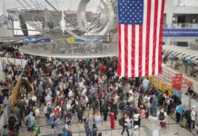 Crowd of travelers waiting in line at an airport security checkpoint under an American flag
