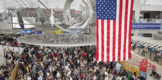 Crowd of travelers waiting in line at an airport security checkpoint under an American flag
