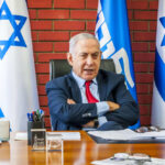 A political leader seated at a desk with Israeli flags in the background