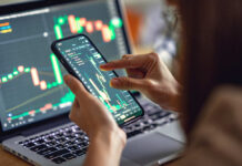 Person analyzing stock market data on a smartphone with a laptop in the background