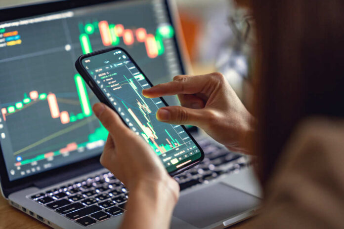 shutterstock_1990259150.jpg Person analyzing stock market data on a smartphone with a laptop in the background