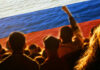 Crowd of people with raised fists in front of a large Russian flag over a water background