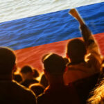 Crowd of people with raised fists in front of a large Russian flag over a water background