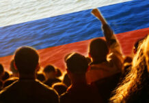 Crowd of people with raised fists in front of a large Russian flag over a water background