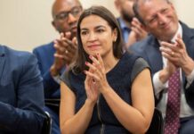 AOC’s 2028 Ambitions: President or Senate? A woman in a dark dress clapping at a political event with an engaged audience in the background