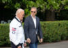 Two men walking outdoors, one in a jacket with an American flag, the other in a blazer