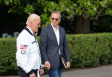 Two men walking outdoors, one in a jacket with an American flag, the other in a blazer