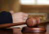 A wooden gavel on a desk with a blurred hand in the background reviewing documents
