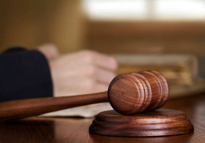 A wooden gavel on a desk with a blurred hand in the background reviewing documents
