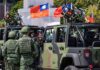 Soldiers in camouflage uniforms standing near military vehicles with flags in the background
