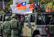 Soldiers in camouflage uniforms standing near military vehicles with flags in the background