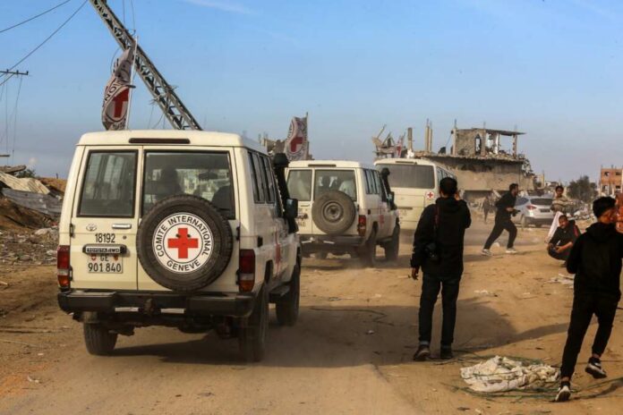 Red Cross vehicles drive through a dusty street with damaged buildings