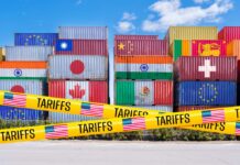 Stacked shipping containers with national flags behind yellow tariffs tape