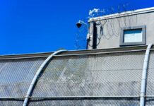 Louisiana Detention Alarms Intensify Security camera mounted above a prison fence with barbed wire against a blue sky