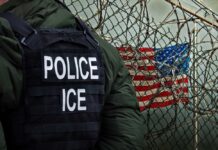 Reporter Calls Armed Ambush A Protest A police officer in a tactical vest stands near a barbed wire fence with an American flag in the background