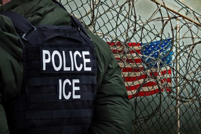 An,Immigration,And,Customs,Enforcement,(ice),Officer,Stands,In,Front A police officer in a tactical vest stands near a barbed wire fence with an American flag in the background