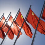 Multiple Chinese flags waving against a clear blue sky with sunlight in the background