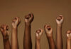 NYC Plays Favorites On Protests A group of diverse hands with raised fists against a brown background