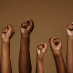 NYC Plays Favorites On Protests A group of diverse hands with raised fists against a brown background