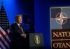 Trump’s NATO Win Comes At A Price A man speaking at a podium with a NATO logo and an American flag in the background