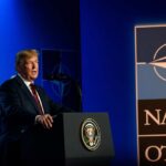 A man speaking at a podium with a NATO logo and an American flag in the background
