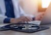 Anti-Fraud Move Hits Medical Suppliers A stethoscope resting on a tablet with a blurred medical professional in the background typing on a laptop