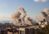 Toxic Skies Return — Who’s Protecting Anyone? Smoke rising from buildings in a city with a coastal view