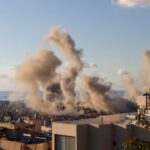 Toxic Skies Return — Who’s Protecting Anyone? Smoke rising from buildings in a city with a coastal view