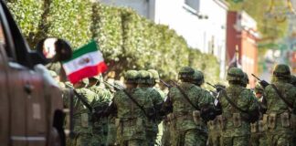 Soldiers in green camouflage march carrying rifles and a Mexican flag