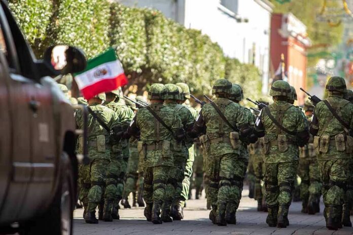 Soldiers in green camouflage march carrying rifles and a Mexican flag