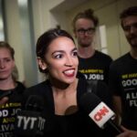 Dem Power Brokers Panic Over AOC Woman speaking to reporters with microphones surrounded by supporters
