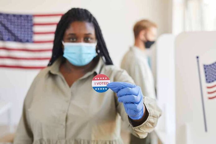 shutterstock_1810359373 Voter wearing a mask holds an I Voted sticker at a polling place