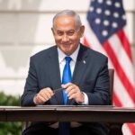 man in a suit sitting at a table with flags in the background