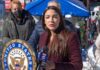 Woman speaking at outdoor press event with microphones and seal