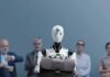 Robot Takeover Slashes Wages—Hotel Grins Humanoid robot holding a briefcase among seated job applicants