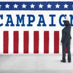 Anti-Corp Crusader’s Donors Raise Eyebrows Man in suit writing on large campaign banner with stars and stripes