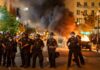 Christian Jones and Teddy Williams among police line at night protest with fire and smoke